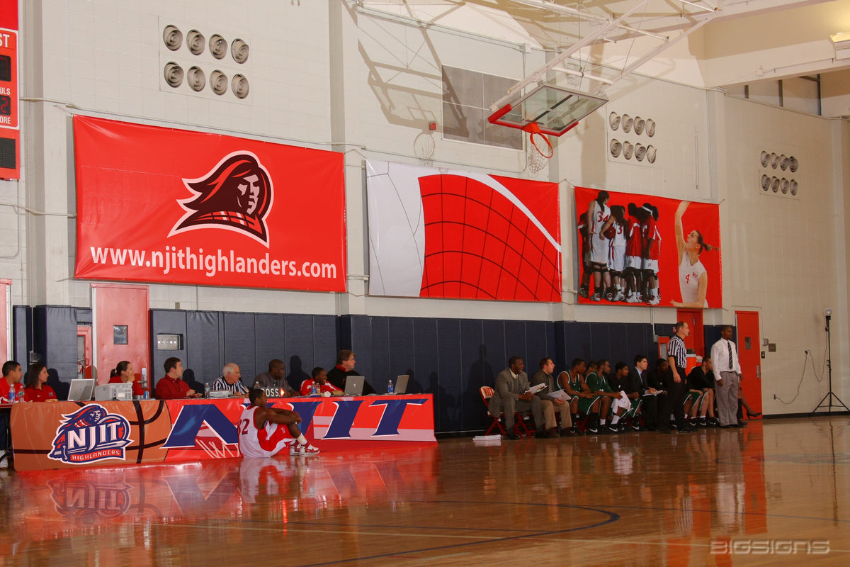 NJIT Highlanders logo and player photo banners on a gym wall above the scorer's table