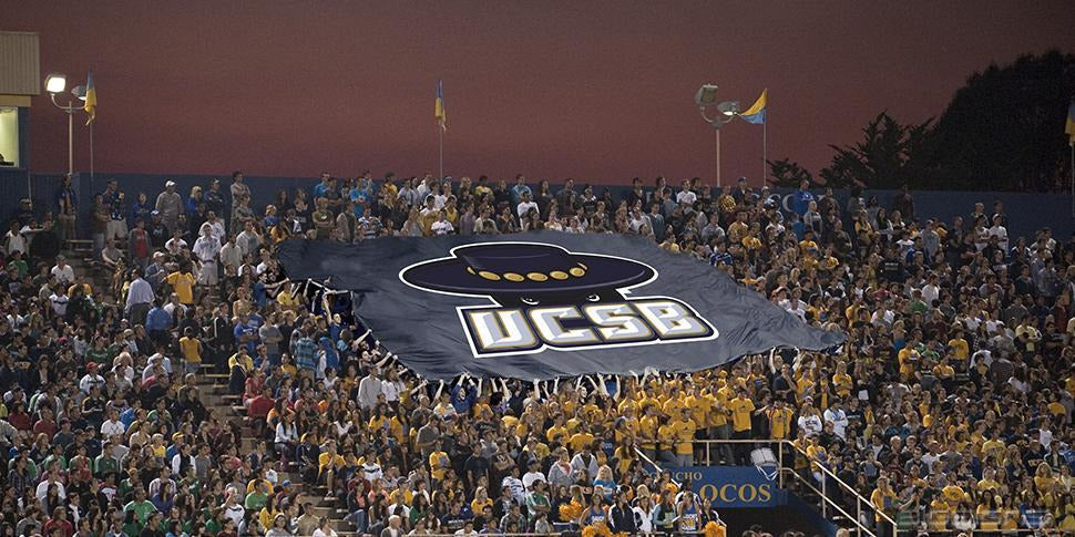 Giant navy UCSB MonsterFlag™ section banner passed over a crowd of fans in the stands at night