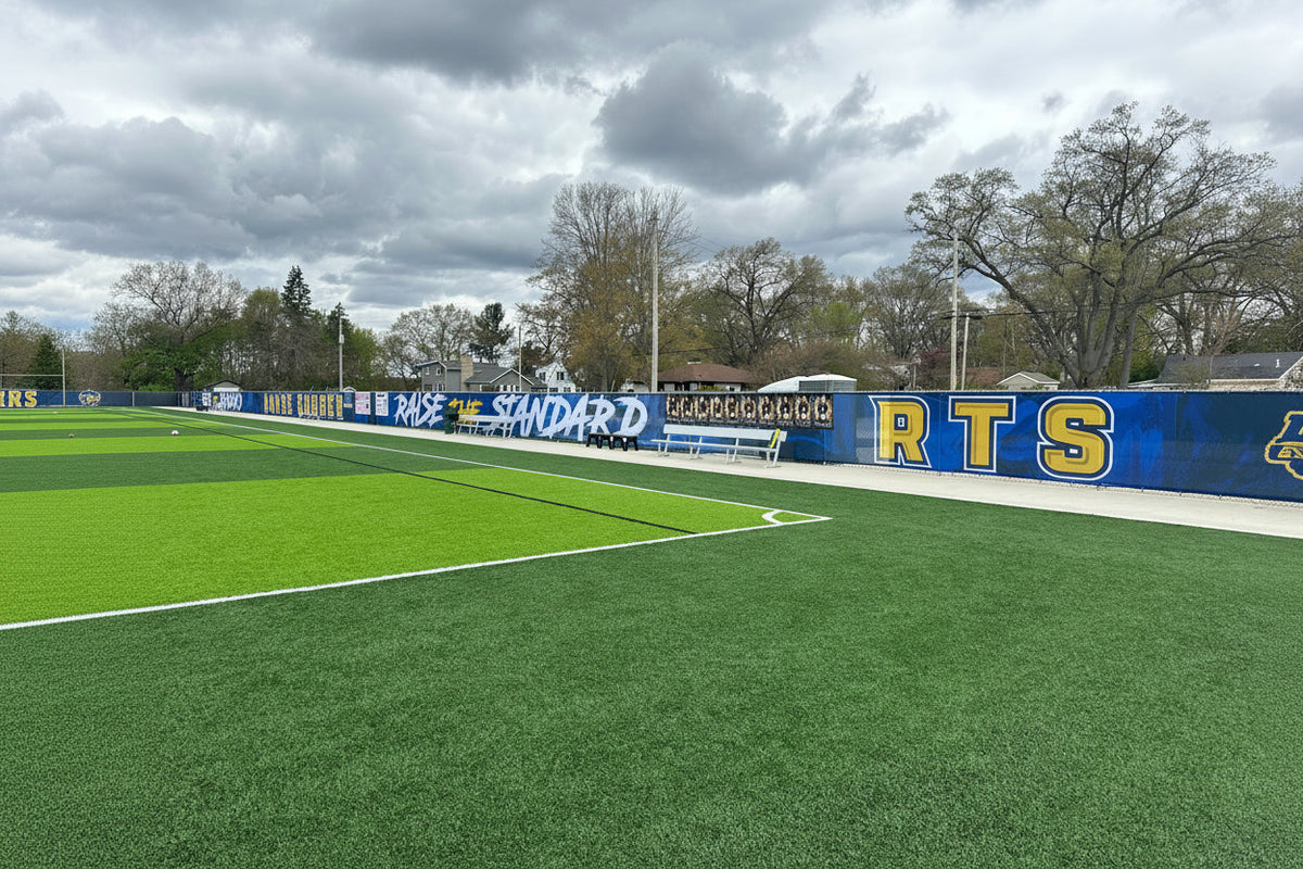 Black mesh athletic windscreen installed along a chain link fence at a school sports facility outfield perimeter