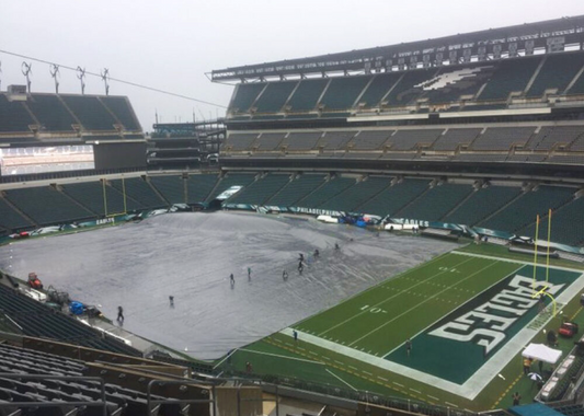 Stadium ground crew deploying a custom waterproof rain tarp across a football field to keep it dry and playable