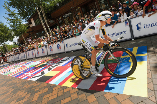 Cyclist crossing a colorful multi-flag pavement decal graphic at a crowded outdoor race event with sponsor banners
