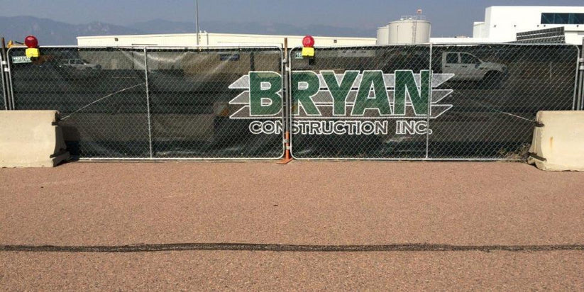 A construction fence screen with logo displaying “Bryan Construction Inc.” attached to a chain-link barrier at the jobsite.