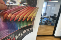Printed DuraMesh sample with colorful feather graphic held by hand, showing perforated mesh banner material in an office.