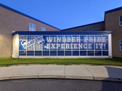 Custom mesh fence windscreen featuring a "Windber" graphic and blue branding on a sports facility chain-link fence.