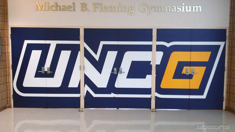 Gym doors wrapped edge to edge in a UNCG wordmark below the Michael B. Fleming Gymnasium sign