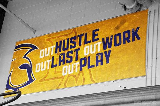 Gold OUTHUSTLE OUTWORK OUTLAST OUTPLAY banner mounted in a Dura-Trac™ track on a block wall