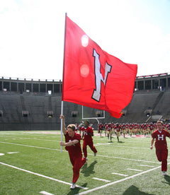 Runner carrying a red flag with a white H onto a stadium field ahead of the team