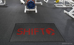 Black LogoRug™ mat with a red Shift logo on rubber flooring in a weight room